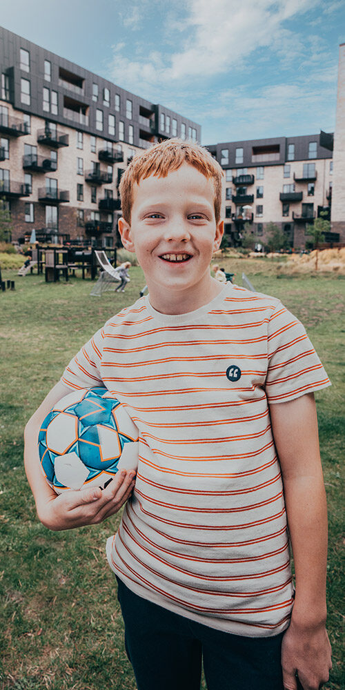A young boy stands with a soccer ball under his arm in front of some houses from Balder.