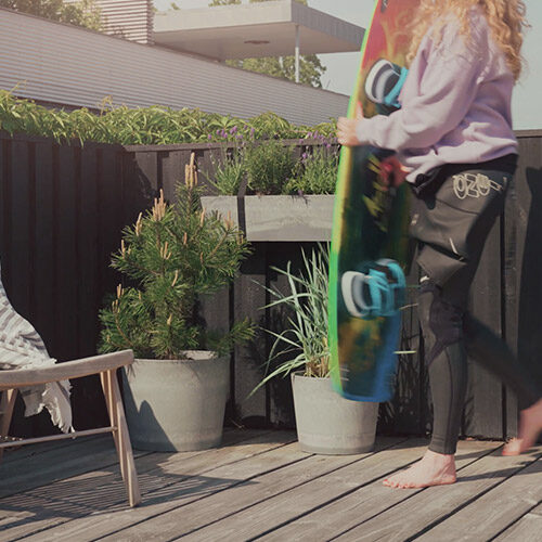 Woman with surfboard walks across terrace. On the terrace there are plants from Greenify.